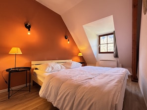 A bedroom with a sloped ceiling, a bed with white bedding, two bedside lamps, and a window with a view of trees. - Joli duplex 5P. Parking & balcon (Cauterets)