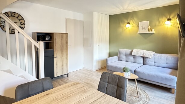 Una sala de estar moderna con una escalera, un sofá gris, una mesa de comedor de madera y una pequeña mesita de noche con una planta.