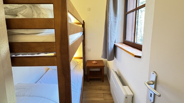 A narrow room with a bunk bed, a small table, a window with curtains, and a radiator.