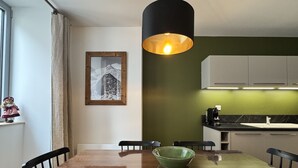 Un comedor con mesa de madera, sillas negras, un tazón verde, lámpara colgante y un cuadro en la pared.