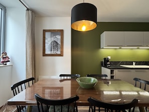 Un coin repas avec une table en bois, des chaises noires, un bol vert, un luminaire suspendu et un tableau encadré au mur.