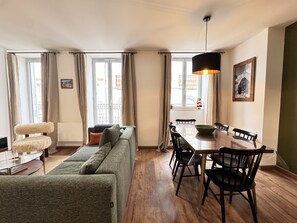 A modern living room with a sofa, dining table, and chairs. There is a window with curtains, a hanging light fixture, and a framed picture on the wall.