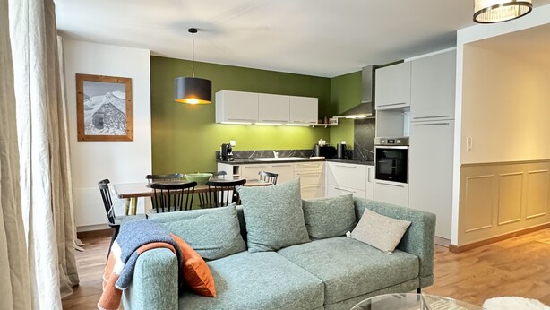 Una sala de estar moderna con un sofá gris, una mesa de centro de vidrio y una zona de comedor con una pared verde y armarios blancos.