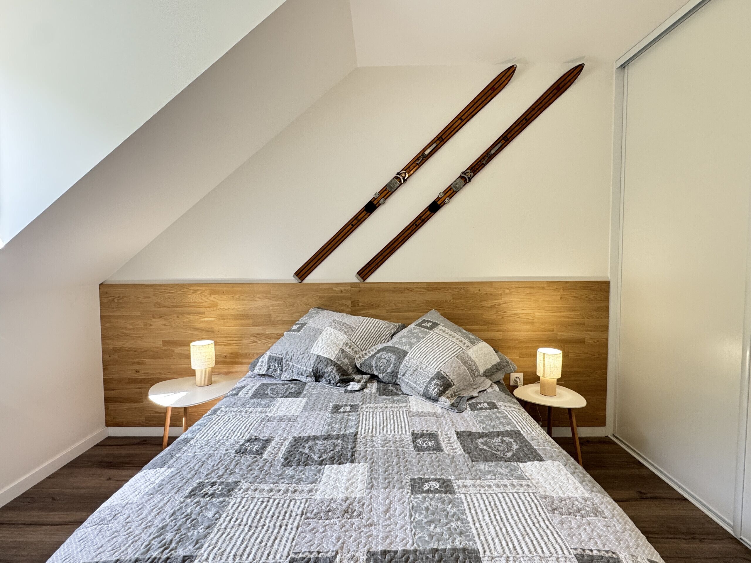 Een slaapkamer met schuine wanden, een houten hoofdbord, een gewatteerde spreideken, twee bedlampen, en twee ski's aan de muur bevestigd.