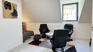 Une petite chambre moderne, équipée d’un lit, de deux chaises noires, d’une petite table et d’une fenêtre laissant entrer la lumière naturelle.