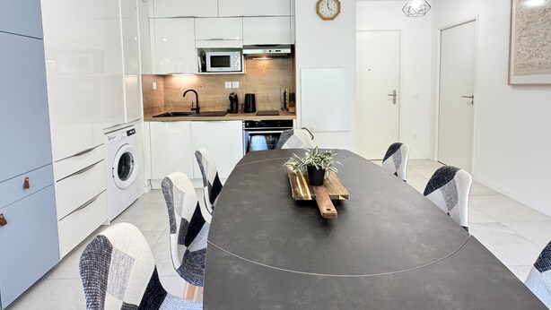 Une cuisine moderne avec des meubles blancs, un four à micro-ondes et un évier. On y trouve une table à manger avec des chaises et un pot de fleurs posé dessus.