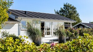 A white house with a dark roof, a covered porch, and a garden with various plants and flowers.