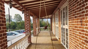 A covered porch with a swing, brick walls, and a view of parked cars. - The Windsor Villa (Trimbak)