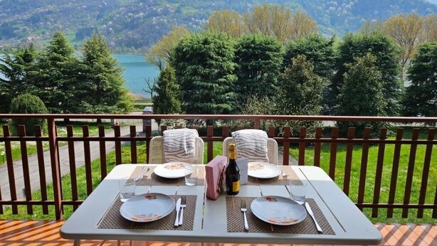 Mesa de comedor para dos personas con una botella de champán, platos y cubiertos, con vistas a un lago y colinas boscosas.