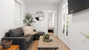 Una sala de estar moderna con un sofá gris, una mesa de centro de madera con objetos decorativos, un espejo en la pared y un televisor montado.