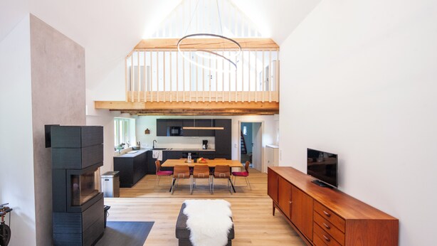 A spacious living area with a high ceiling, wooden beams, a fireplace, a dining table with chairs, and a television on a wooden cabinet.