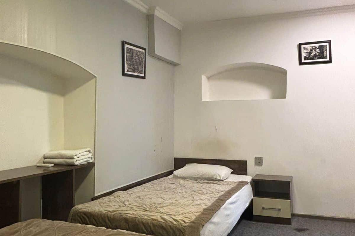 A hotel room with two single beds, a small desk with a chair, a nightstand, and a wall-mounted shelf.