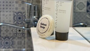 A bathroom with a shower, a mirror, and toiletries including a bottle of Calvados shampoo and a bar of Sabun soap. - Central Budget GH  (Baku)