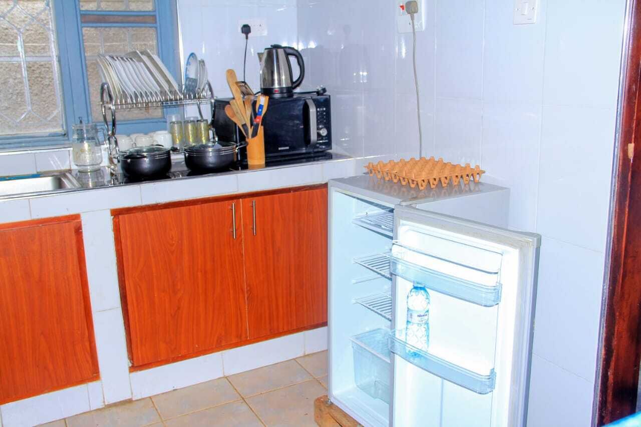 A kitchen with wooden cabinets, a microwave, a kettle, and a refrigerator with a water bottle inside.