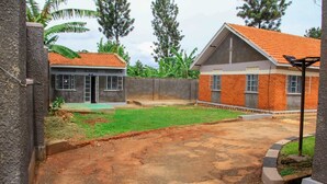 A residential area with a paved driveway, a grassy lawn, and two buildings with brick and concrete exteriors. - Comfortable Private Home in the Heart of Jinja City -Tenzi BnB with WIFI (Jinja)