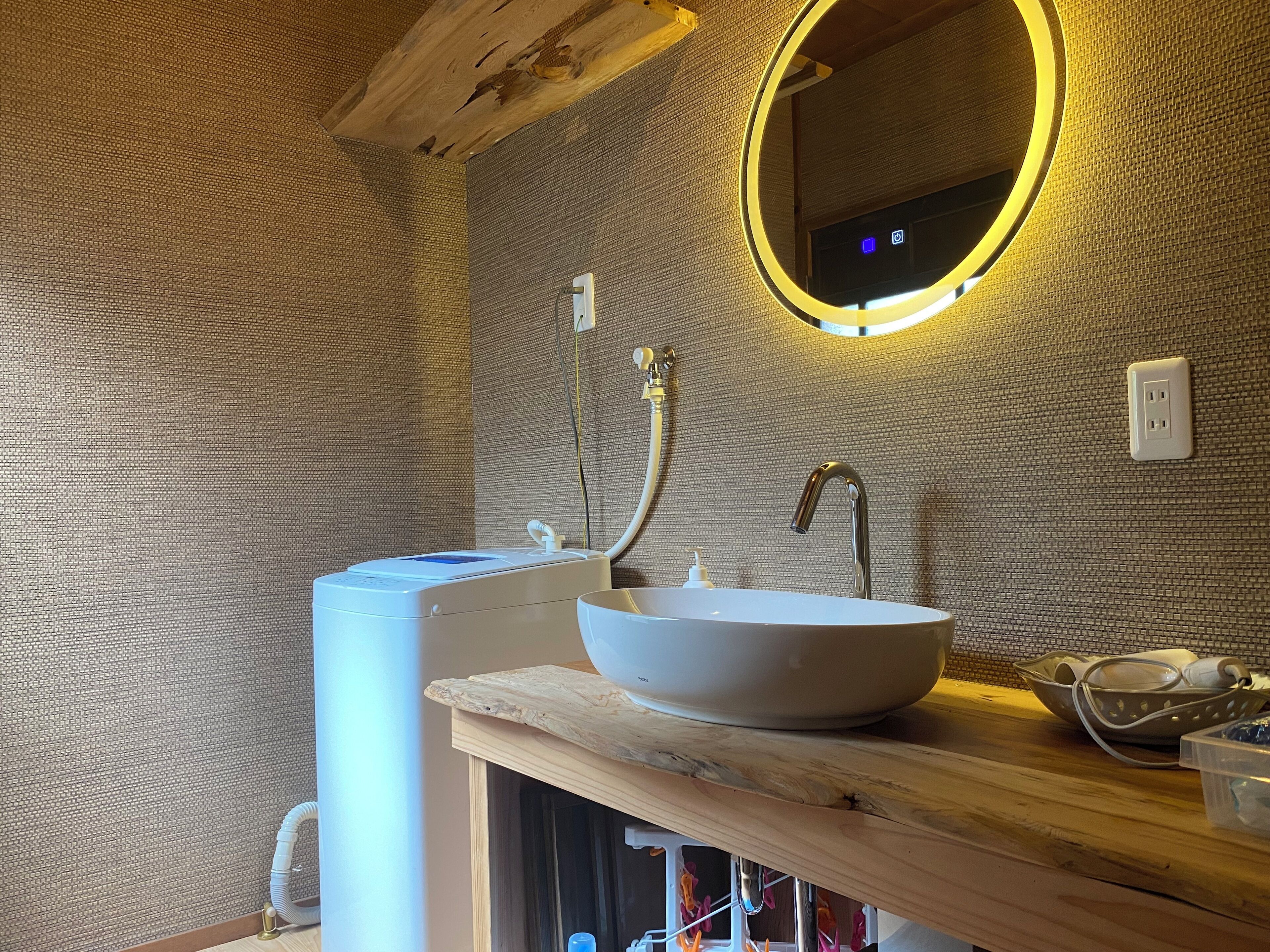 Een badkamer met een wit wastafel, een houten aanrechtblad, een spiegel met rondom verlichting en een structuurwand.