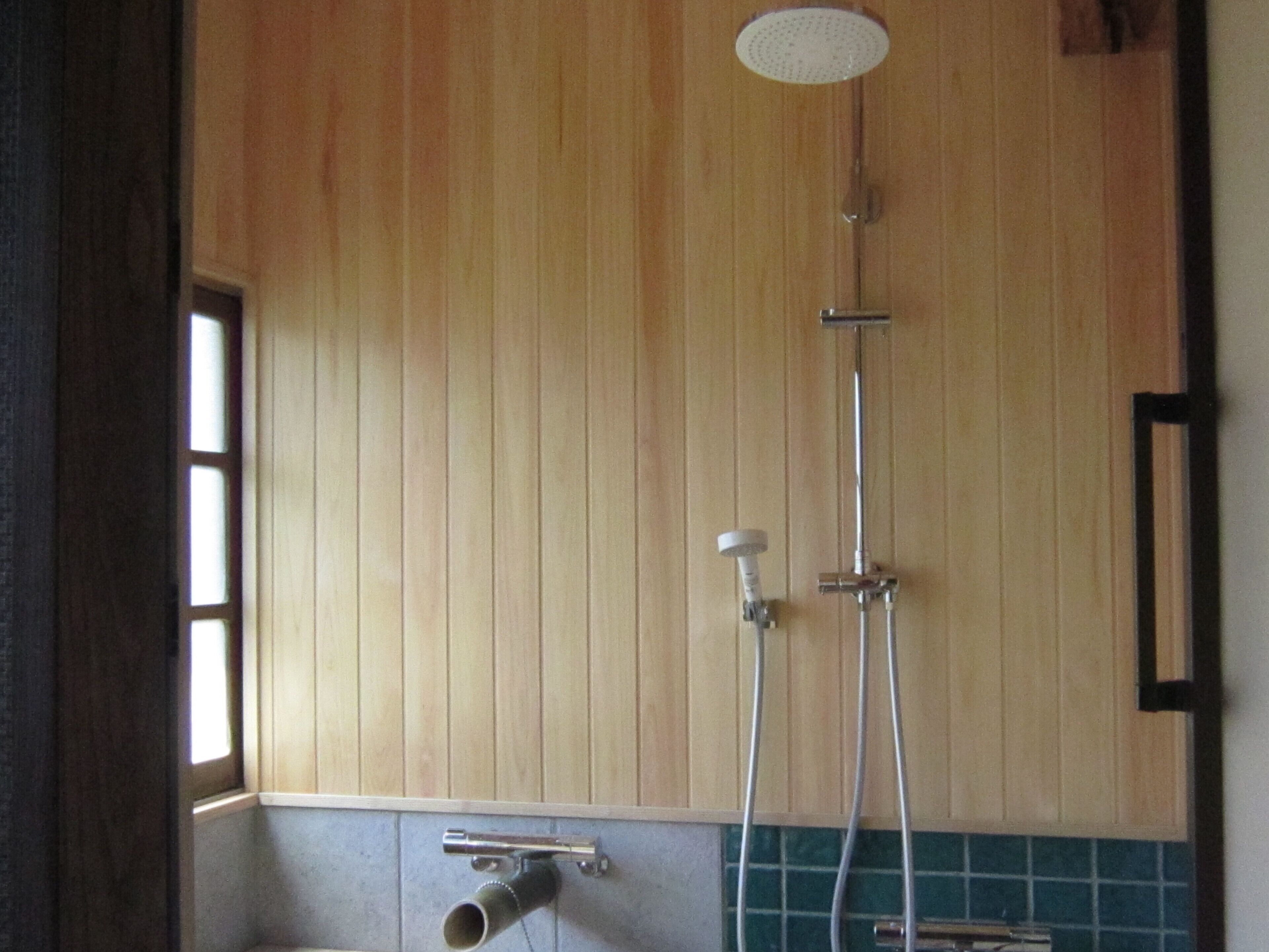A shower with a handheld showerhead and a wall-mounted faucet, wooden paneling, and a window on the left.