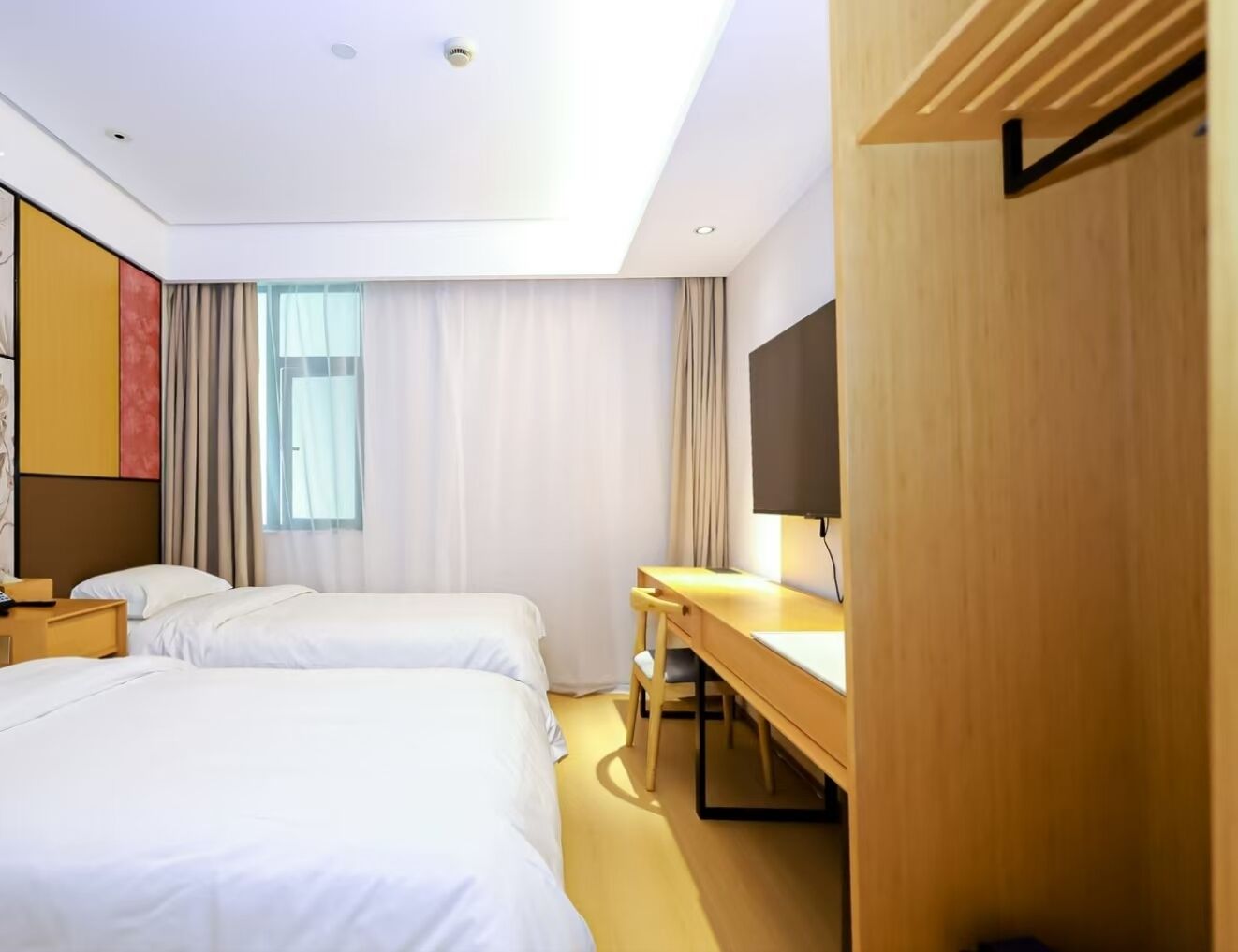 A hotel room with two beds, a desk, a chair, a flat-screen TV, and a window with curtains.