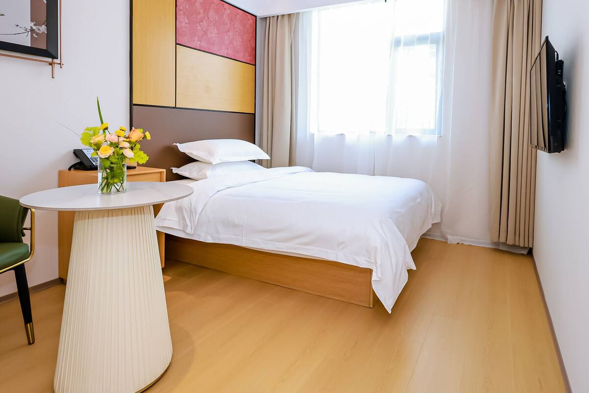 Quarto de hotel com cama, mesa de cabeceira com um vaso de flores, uma cadeira verde e uma televisão na parede.