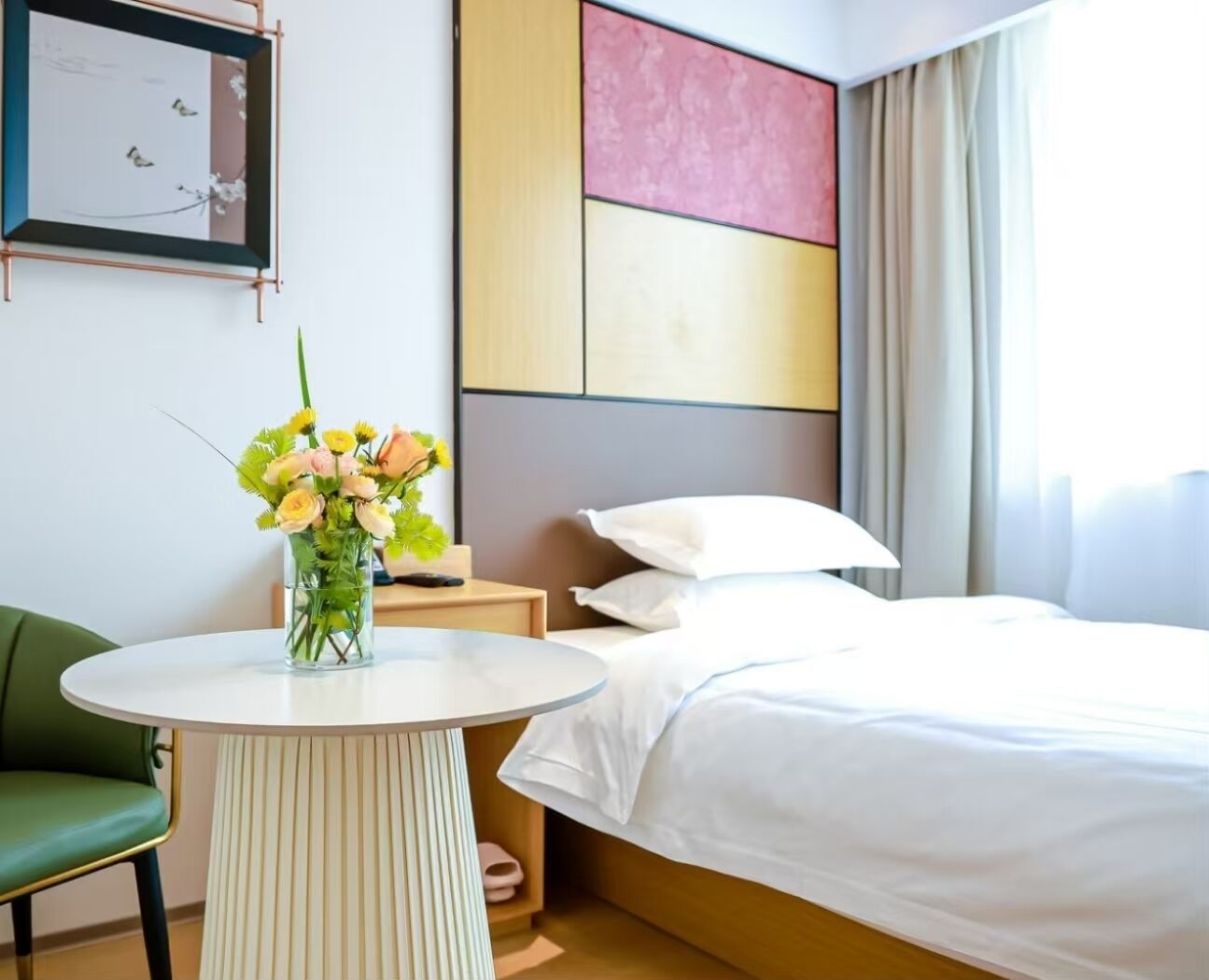 Habitación de hotel con cama, una mesita de noche con un florero y una silla verde, además de un cuadro en la pared.
