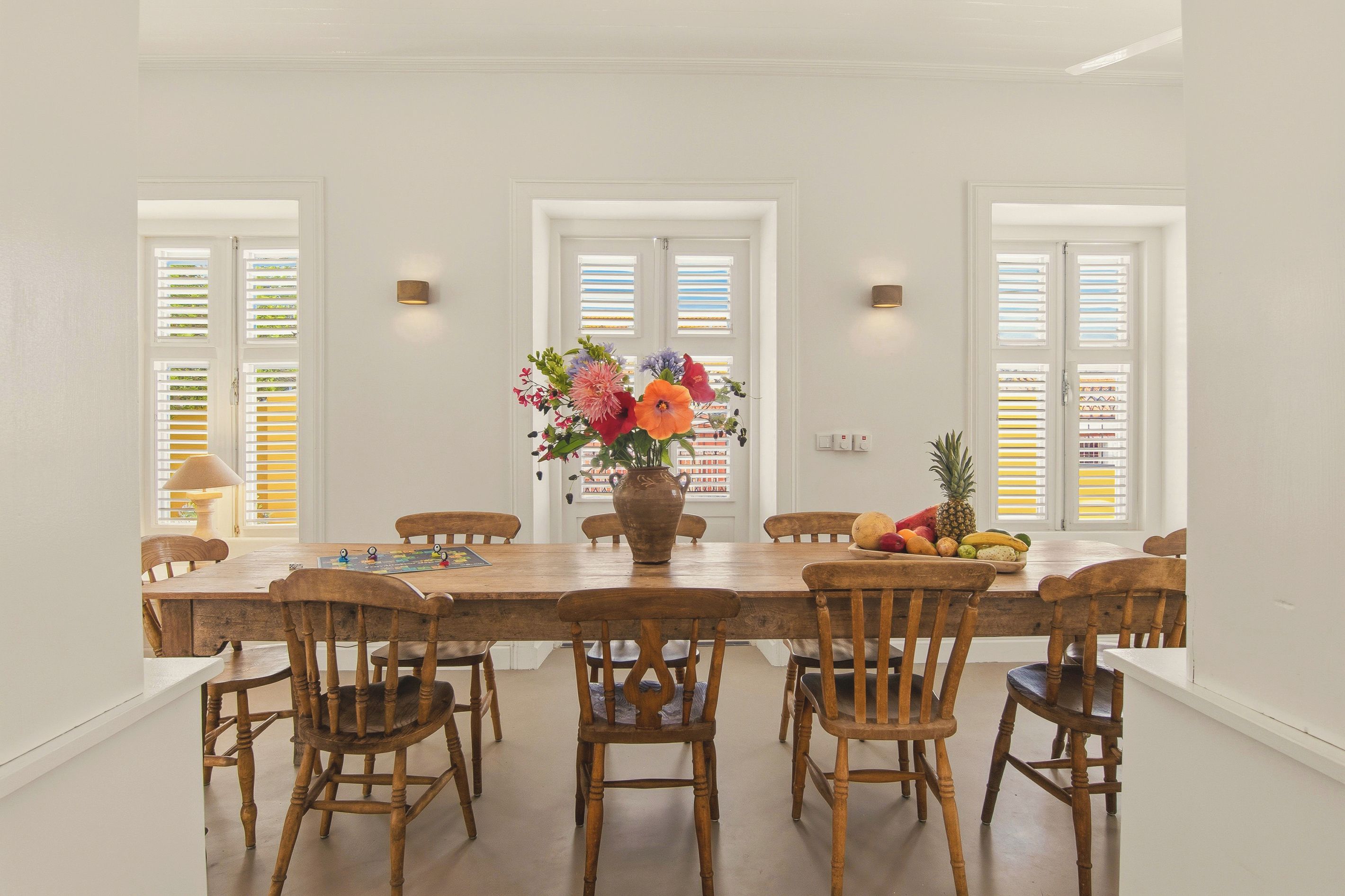 Une salle à manger avec une table et des chaises en bois, un vase rempli de fleurs et un panier de fruits.