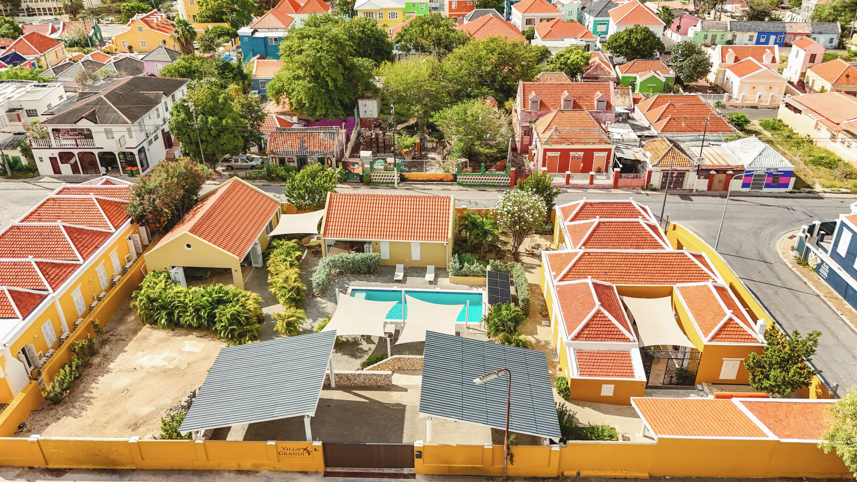 Un quartier résidentiel avec des maisons aux couleurs vives, une piscine et une entrée sécurisée par un portail.
