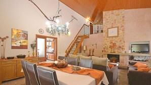 Uma sala de jantar com mesa posta para quatro pessoas, uma escadaria, uma sala de estar com lareira e uma televisão.