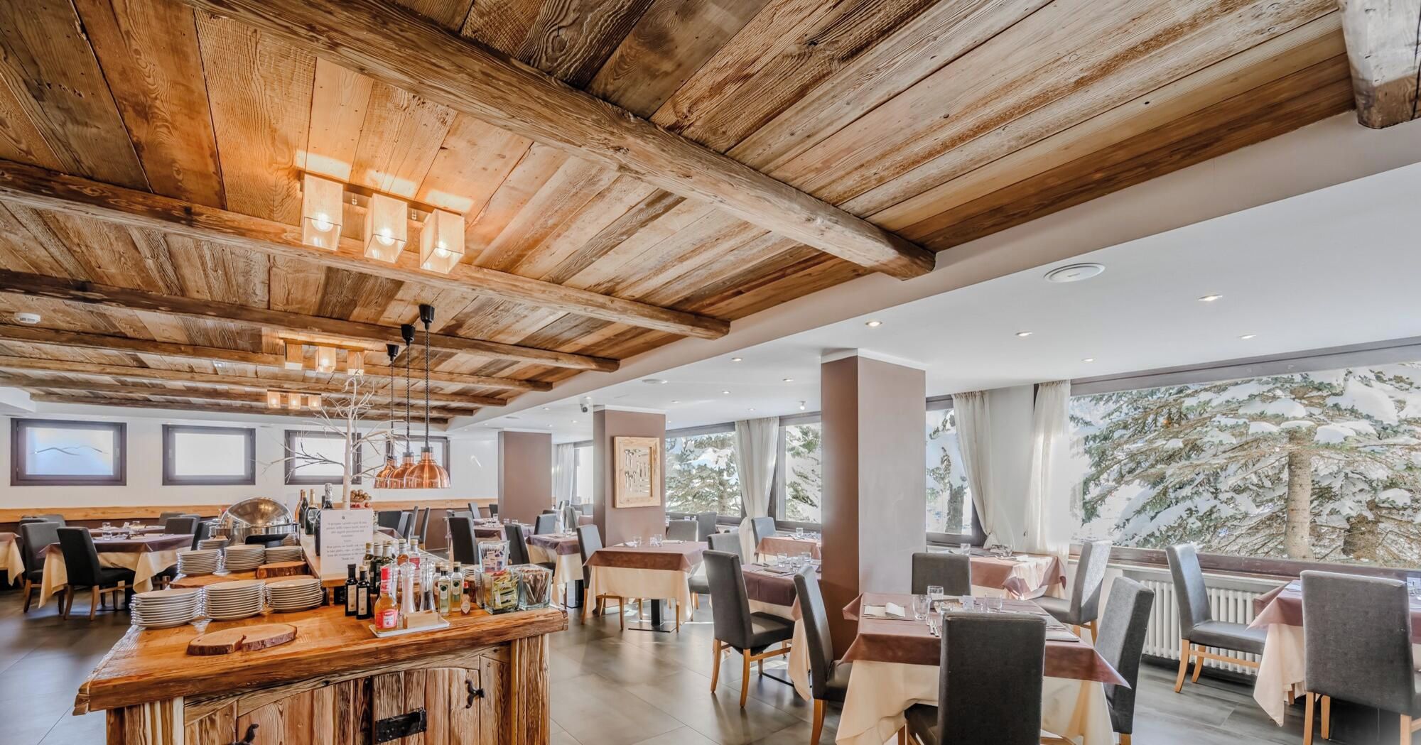 Un restaurant spacieux avec des plafonds en bois et un bar central. Les tables sont disposées pour accueillir les clients, et de grandes fenêtres offrent une vue sur l’extérieur.