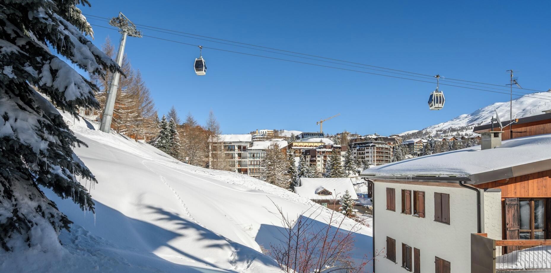 A snow-covered ski slope with a cable car system, buildings, and a clear blue sky.