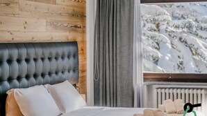 A bedroom with a tufted headboard, a bed with white linens, a window with a view of snow-covered trees, and a radiator. - Hotiday Sestriere Ski Run (Sestriere)