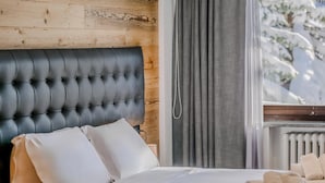 Un dormitorio con cabecera capitonada, ropa de cama blanca y vistas a árboles cubiertos de nieve a través de una ventana.