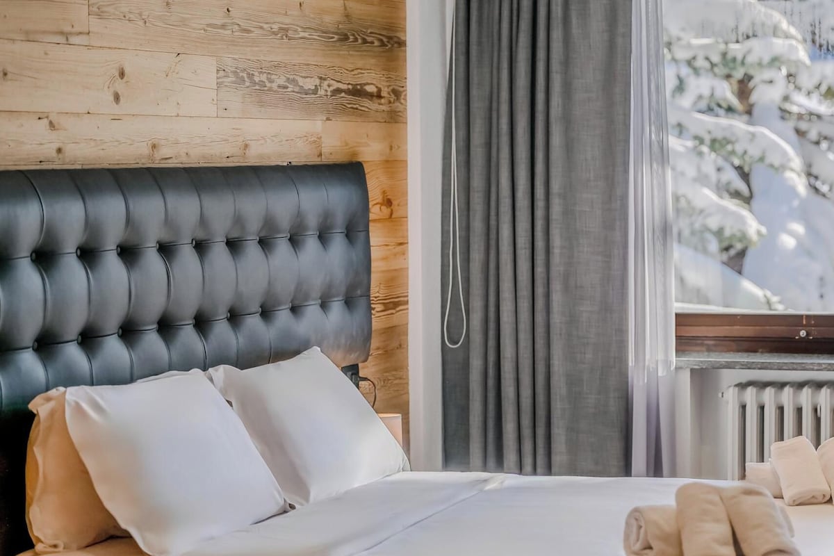 A bedroom with a tufted headboard, a bed with white linens, a window with a view of snow-covered trees, and a radiator.