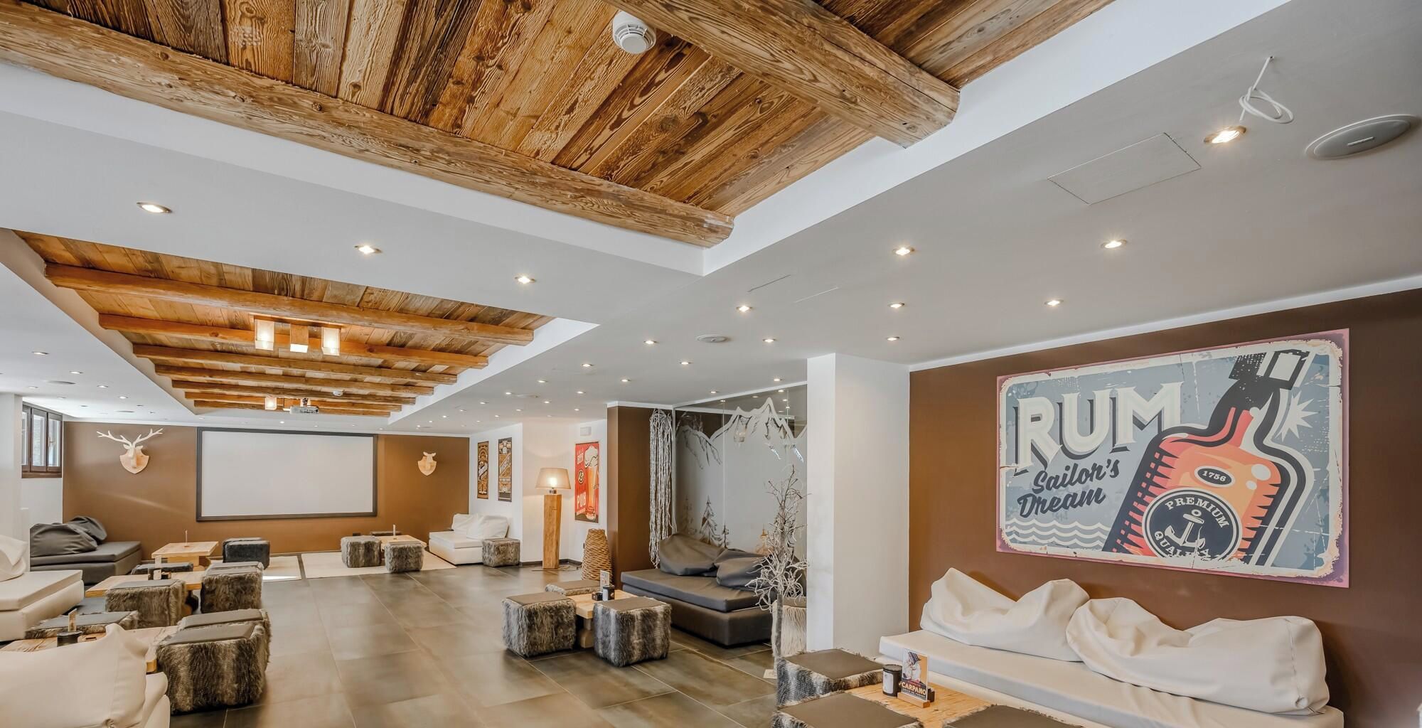 A spacious lounge with a high wooden ceiling, comfortable seating, a large screen, and a wall mural.