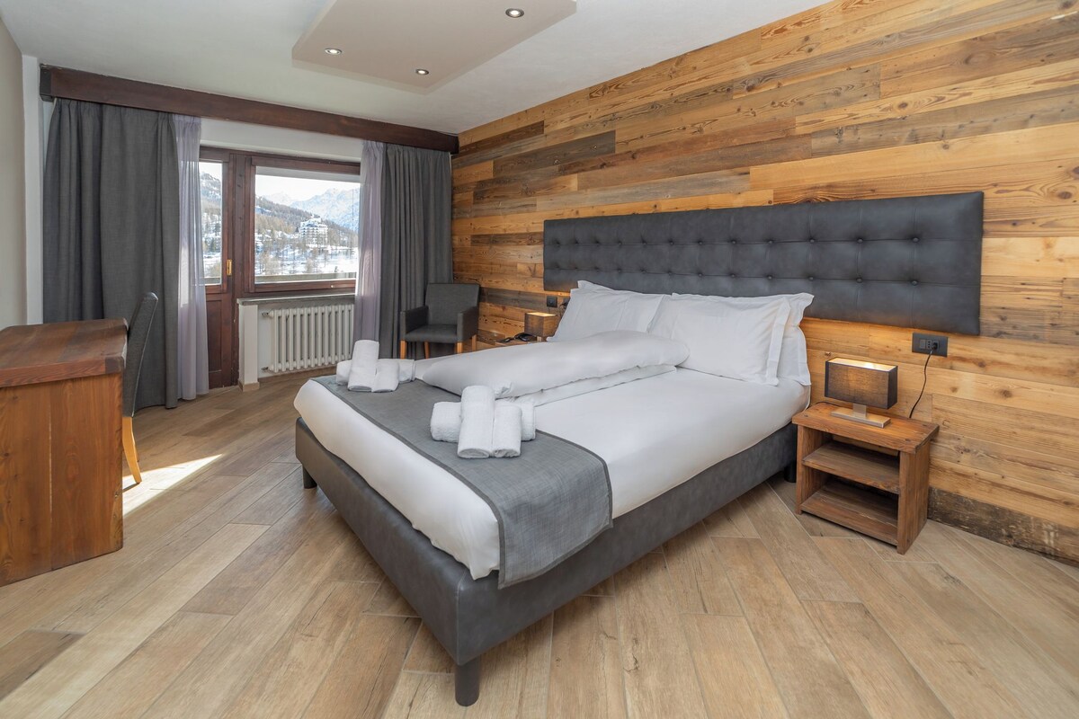A modern hotel room with a large bed, wooden panel walls, a view of mountains, and a small desk.