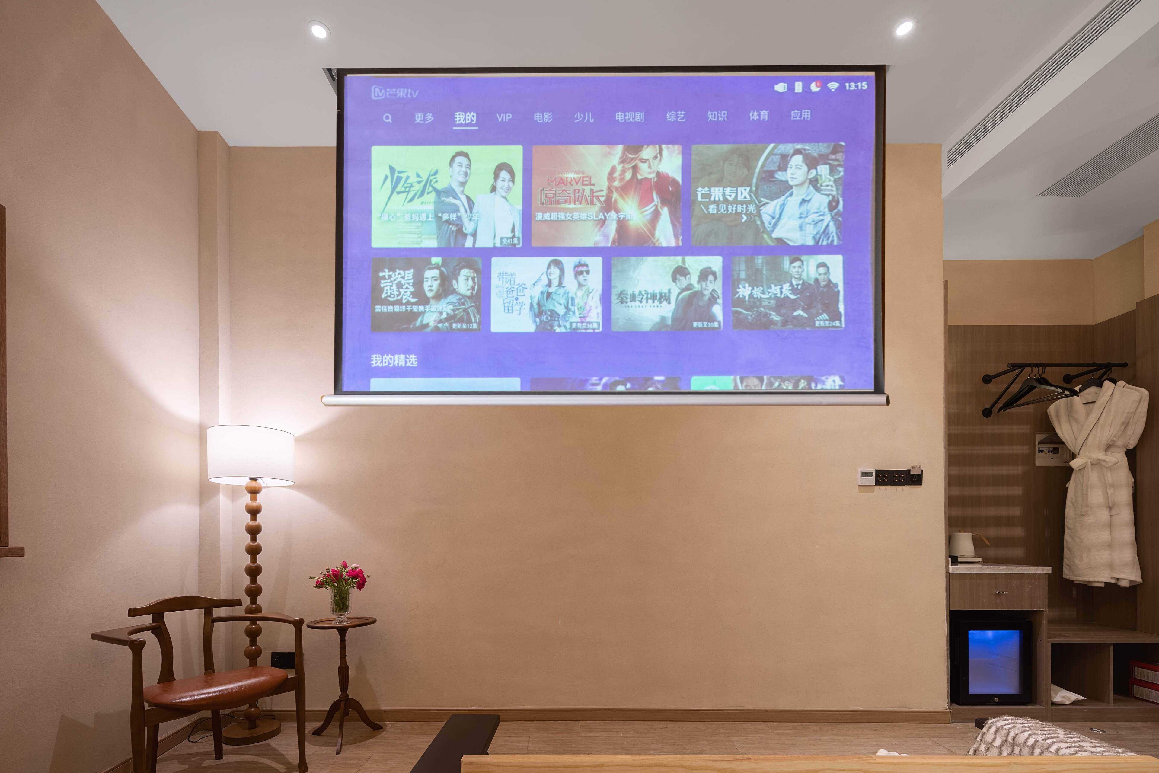 Una habitación con un televisor de pared mostrando carteles de películas, una silla de madera, una mesita con un jarrón y una bata colgada en un perchero.