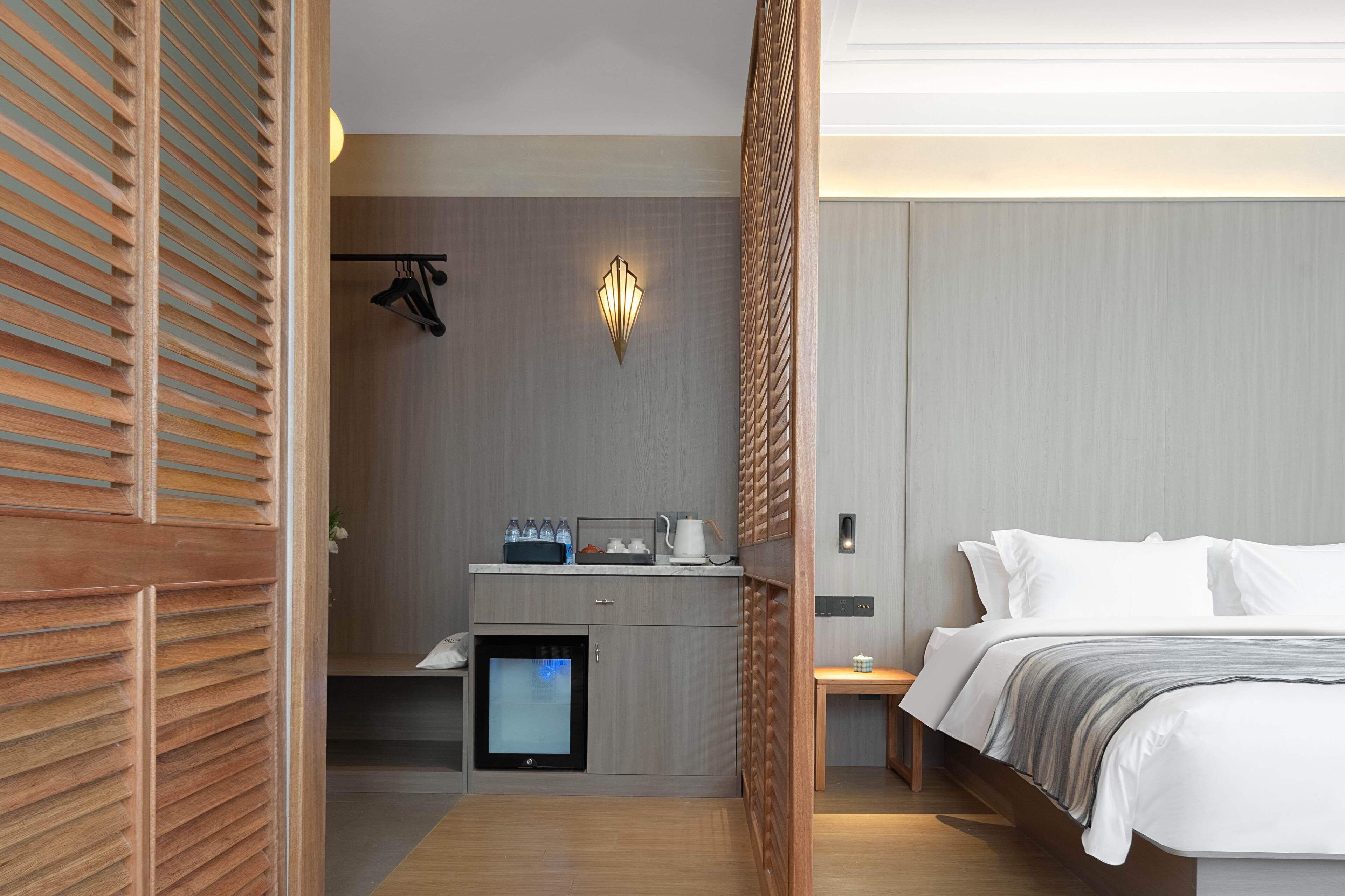 A modern hotel room with a wooden partition, a kitchenette, a bed with white linens, and a wall-mounted light fixture.