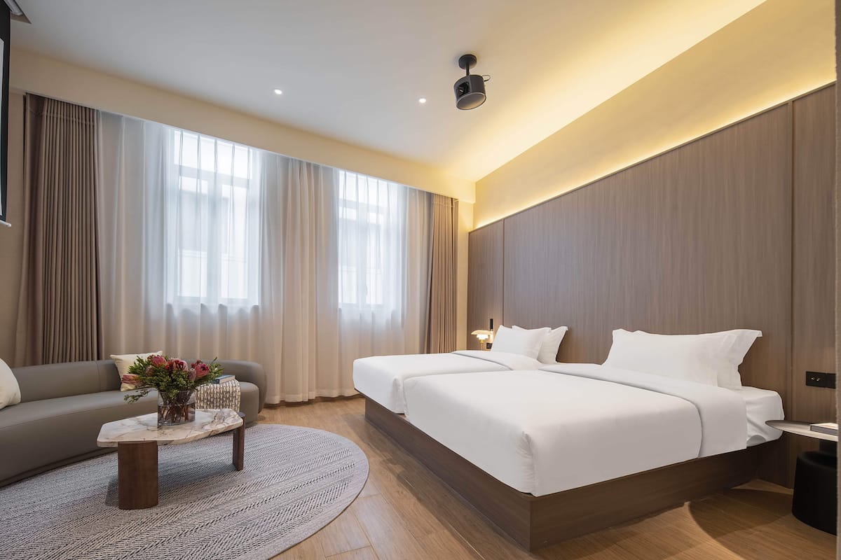 A hotel room with a large bed, a sofa, a coffee table with flowers, and a view of the outdoors through large windows.