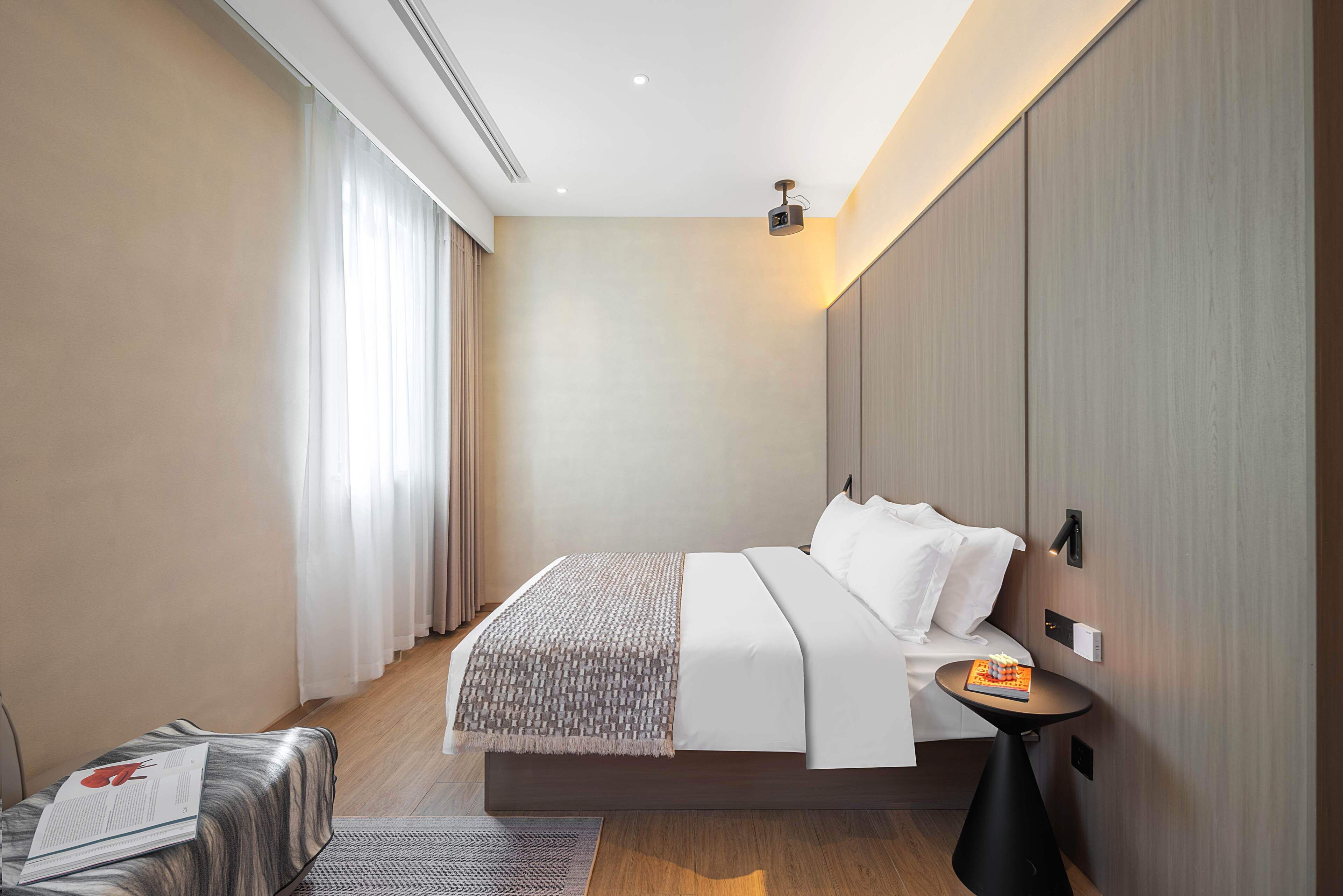 Ein modernes Hotelzimmer mit einem großen Bett, einem Nachttisch mit einem Buch und einem an der Wand montierten Projektor.