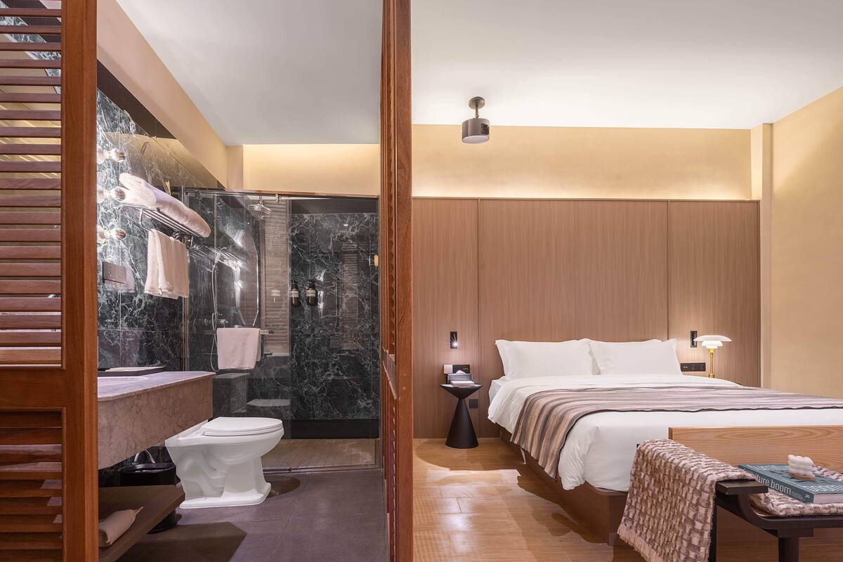 A modern hotel room with a marble bathroom, wooden paneling, a large bed, and a bedside table with a book.