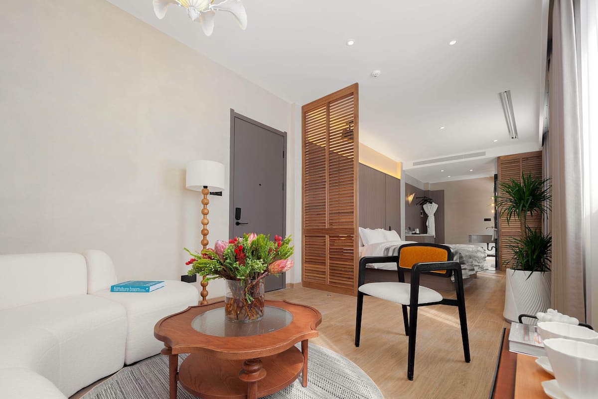 A modern hotel room with a white sofa, a wooden dining table with a vase of flowers, a dining area with a chair, and a bed with a wooden headboard.