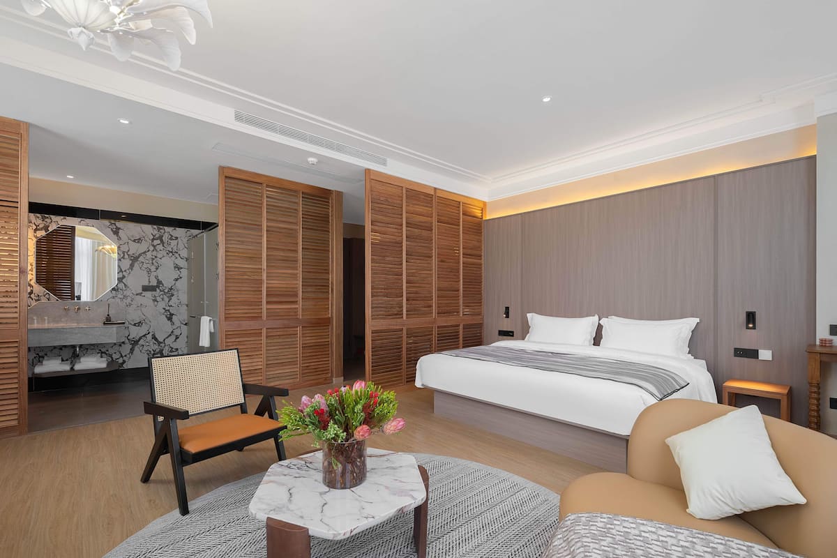 A modern hotel room with a large bed, wooden closet, marble bathroom, and a seating area with a vase of flowers.