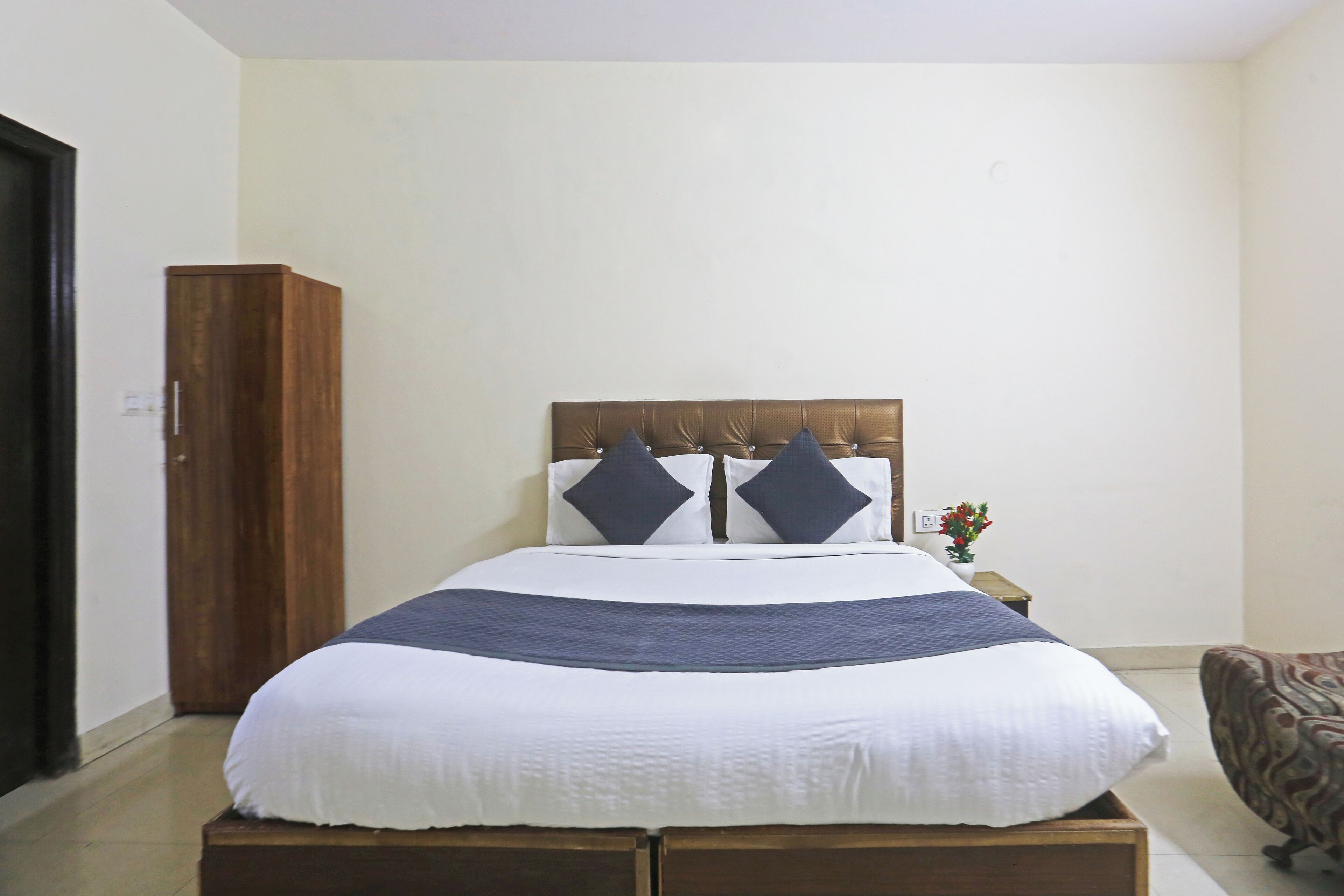 A neatly made bed with a wooden headboard, a single nightstand with a vase of flowers, and a patterned armchair.