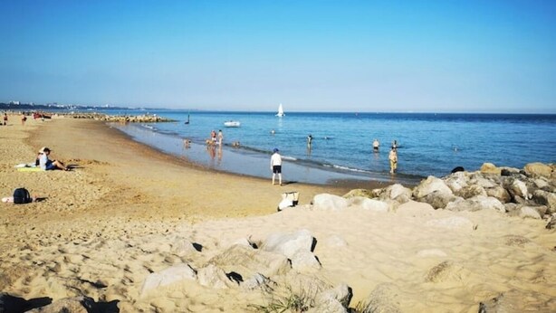 A beach with people swimming and sunbathing, a sailboat in the distance, and a rocky shoreline.