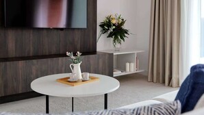 A modern living room with a flat-screen TV mounted on the wall, a round coffee table with a tray, a vase with flowers, and a sofa.