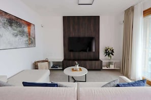 A modern living room with a sofa, a coffee table, a flat-screen TV mounted on a dark wood panel, and a window with curtains.