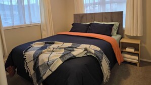 A bedroom with a bed, two windows with curtains, a bedside table with a lamp, and a headboard. - Charming 3-bedroom house in Papakura 20min from Airport (Auckland)