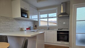 A modern kitchen with white cabinetry, a central island, a built-in oven, and a window providing natural light. - Charming 3-bedroom house in Papakura 20min from Airport (Auckland)
