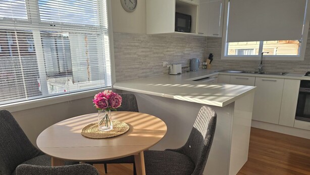 A modern kitchen with white cabinetry, a round wooden table with two chairs, and a vase with flowers. - Charming 3-bedroom house in Papakura 20min from Airport (Auckland)