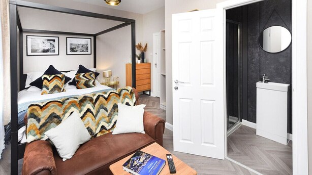 Une chambre à coucher avec un lit à baldaquin, un canapé en cuir marron, une table basse avec un livre et une télécommande, et une porte donnant sur une salle de bain équipée d’un miroir et d’un lavabo.
