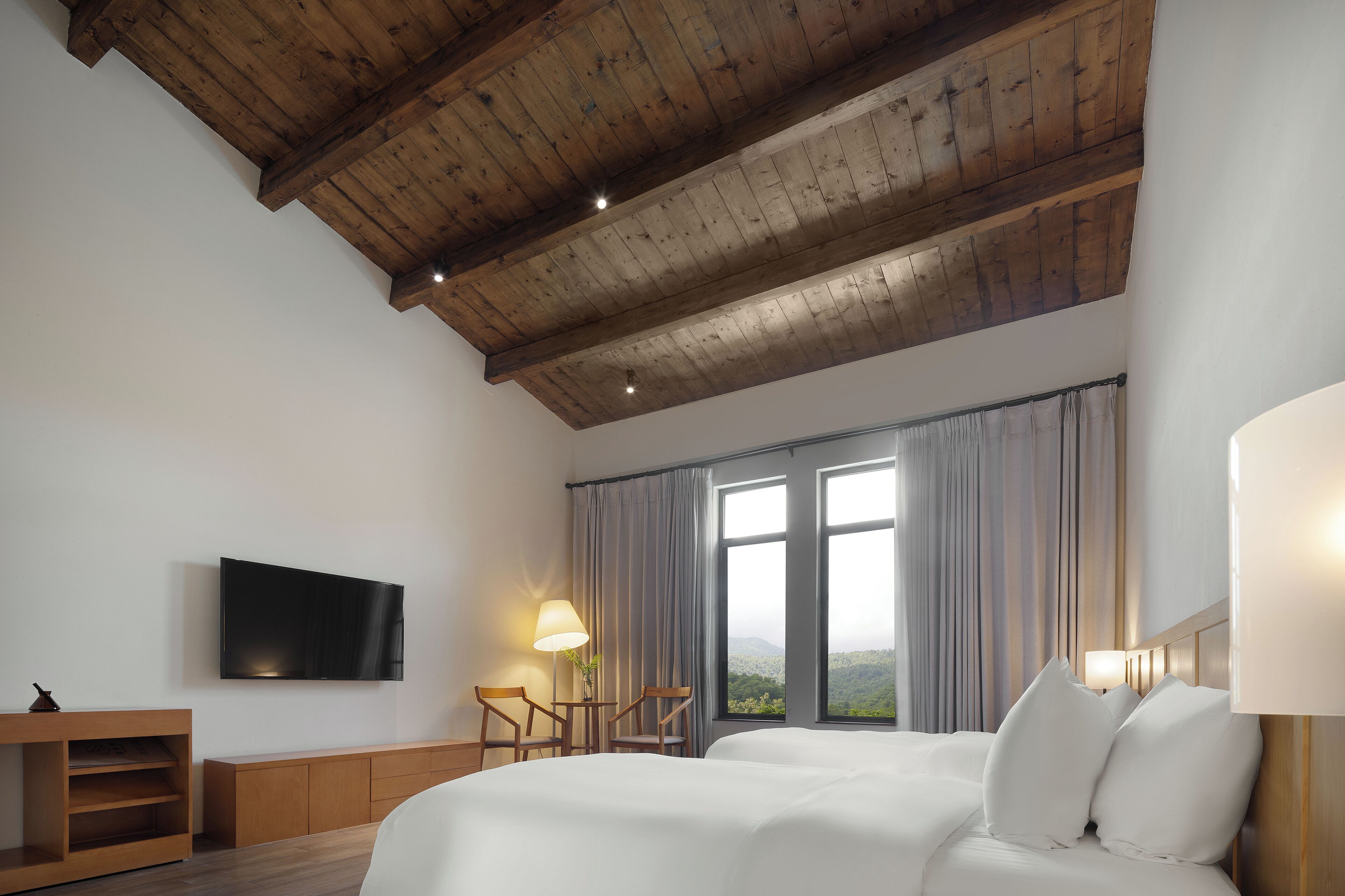 A hotel room with a wooden ceiling, a bed with white bedding, a TV mounted on the wall, and a window with curtains.
